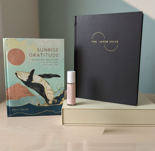 A styled product arrangement on a light wood surface featuring a hardcover book titled 'Sunrise Gratitude' by Emily Silva, a large black notebook or journal with gold lettering reading 'The Inner Voice,' a small pink bottle with a white cap, and a cream-colored box.